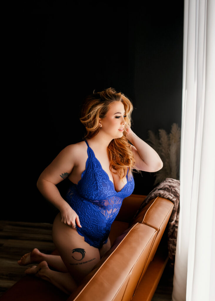 beautiful tattooed full figured woman kneeling on back of couch in blue lace lingerie posing for a boudoir photoshoot in Richmond Virginia.