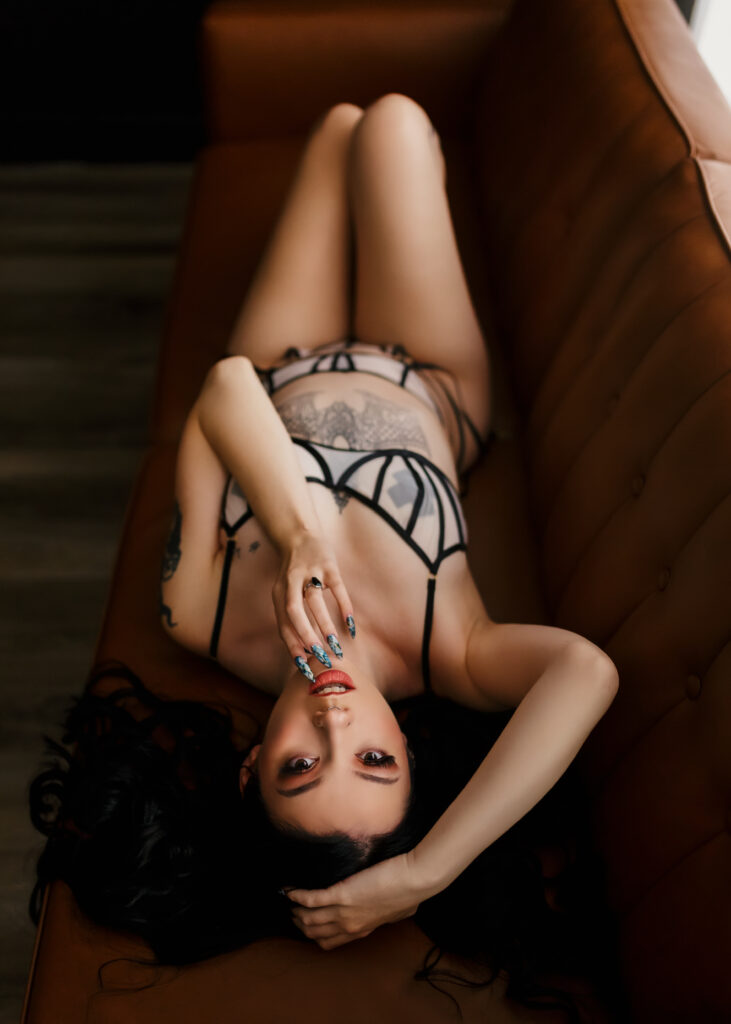 Boudoir client with black hair and tattoos posing on leather sofa in sexy black and nude lingerie. 
