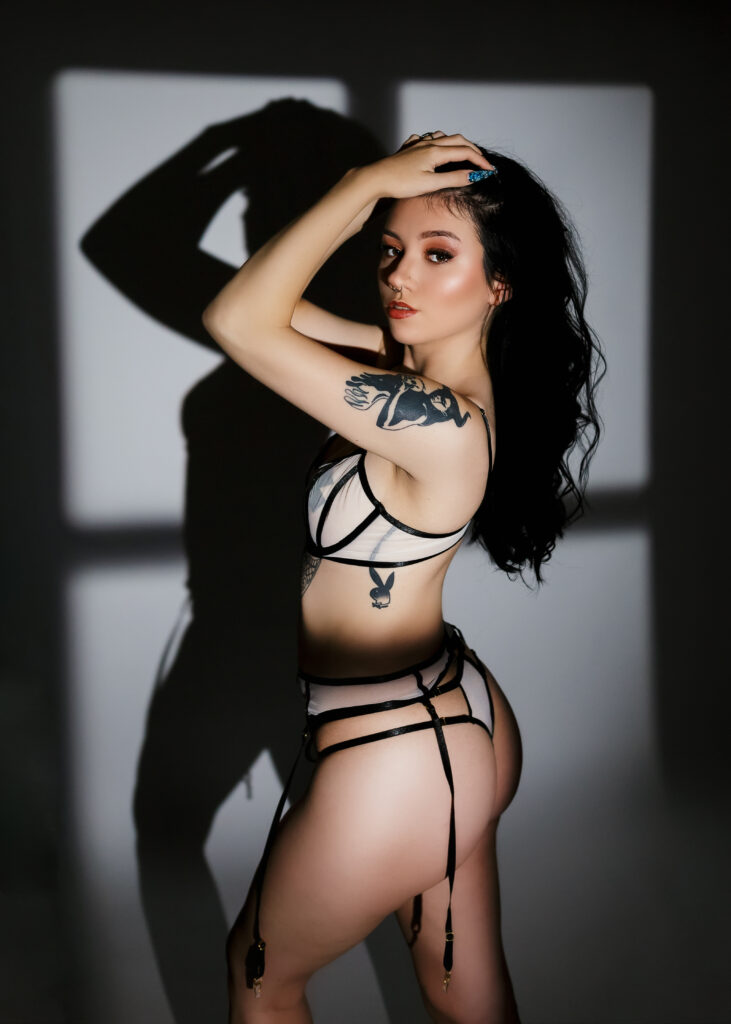 Tattooed woman is posing on a white backdrop with dramatic window light un sext black lingerie. 
