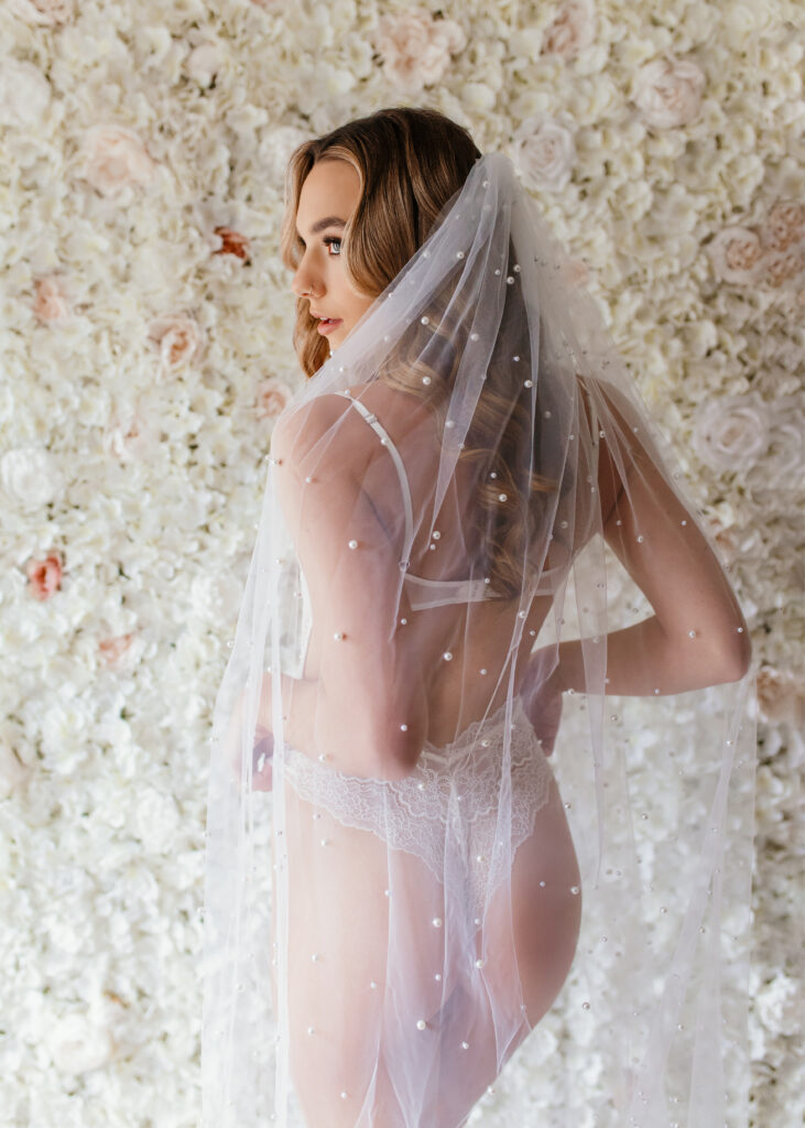 woman is posing in white lingerie with pearl veil in front of flower wall during bridal boudoir photoshoot in Richmond Virginia 