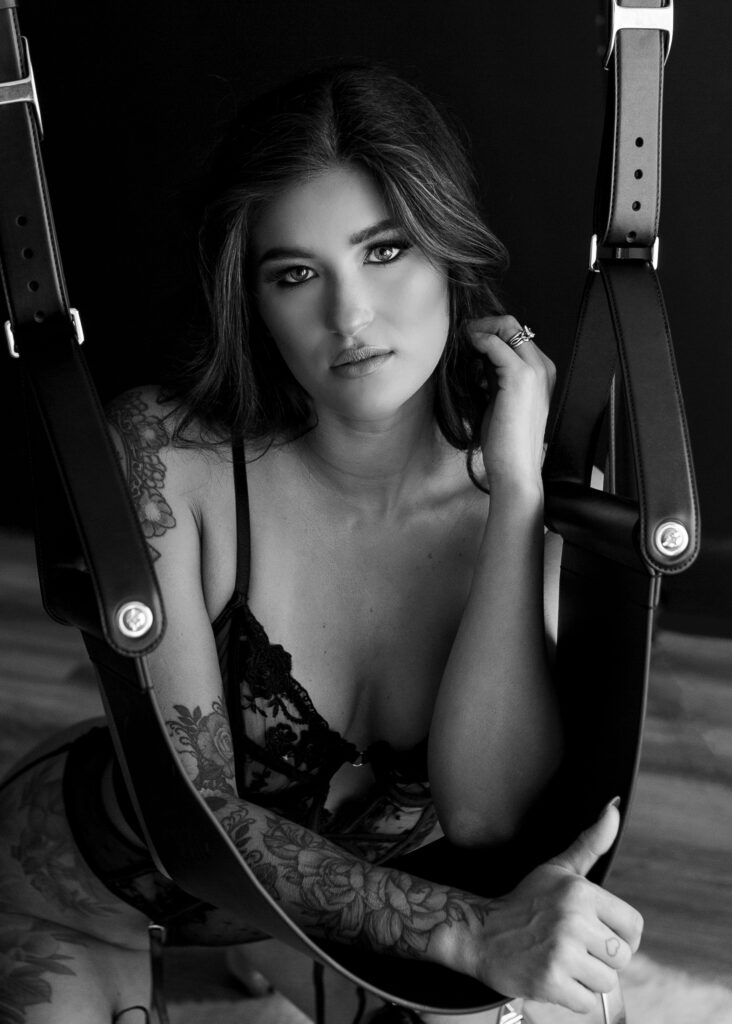Black and white photo of Sexy boudoir client kneeling on floor and posing in front of leather swing. 